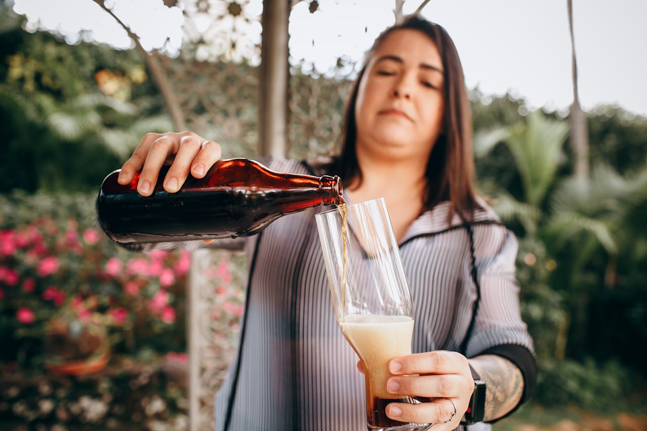 A woman pours craft beer from a bottle into a glass in a lush garden setting.