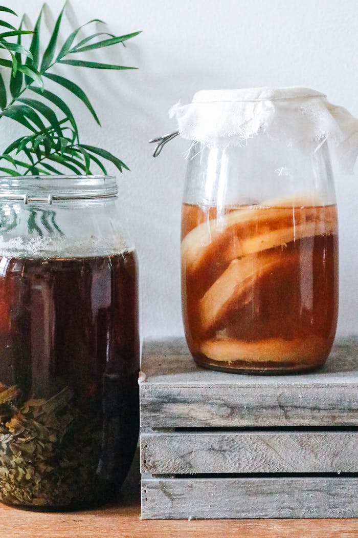 Glass jars with delicious kombucha on wooden board near freshly brewed dark herbal drink in light room