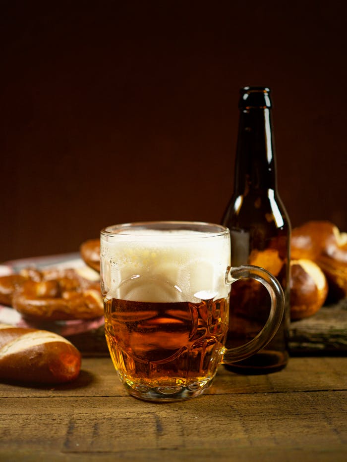 A frothy mug of beer with a bottle on a wooden table, surrounded by fresh bread.